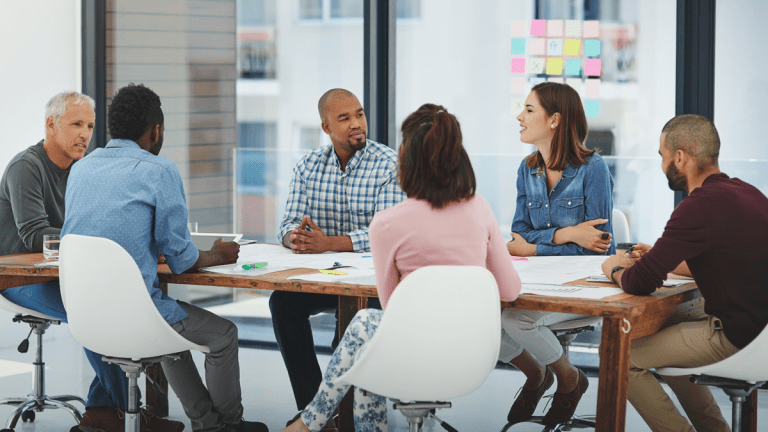 En grupp personer sitter runt ett bord och diskuterar. Bordet är fullt av olika papper. På väggen bakom dem hänger flera postit-lappar.