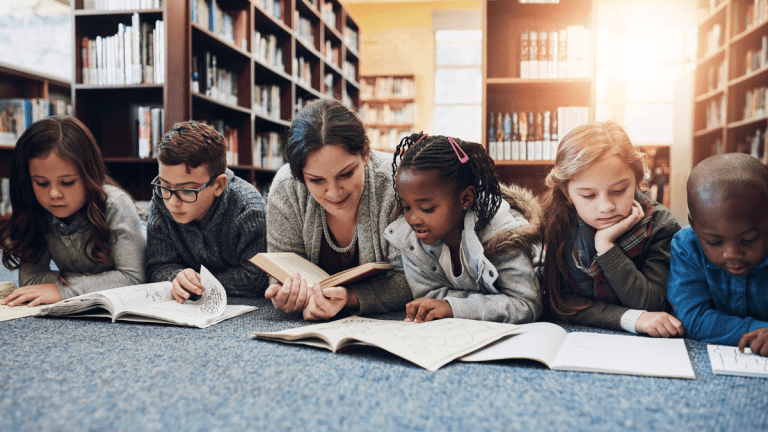 En vuxen kvinna omgiven av fem barn i ett bibliotek. Alla har något läsbart framför sig och ser ut att läsa. De bär stickade och långärmade kläder och ser höstmysiga ut.
