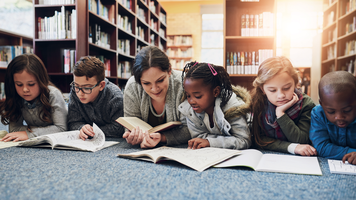 En vuxen kvinna omgiven av fem barn i ett bibliotek. Alla har något läsbart framför sig och ser ut att läsa. De bär stickade och långärmade kläder och ser höstmysiga ut.