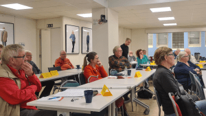 Flera personer sitter i lokalen Åttiotvåan. De har gula namnlappar framför sig på borden. Alla ser framåt mot den som talar längst fram i lokalen.