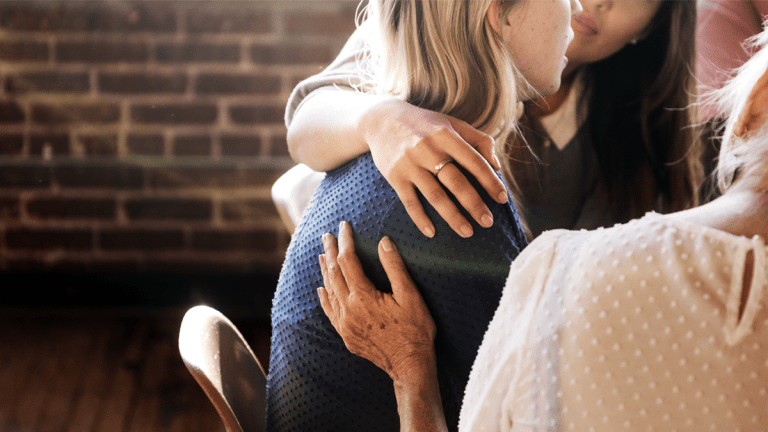 Tre kvinnor sitter tillsammans. Kvinnan till vänster har lagt armen om axlarna på kvinnan i mitten. Kvinnan till höger håller handen på mittenkvinnans rygg.