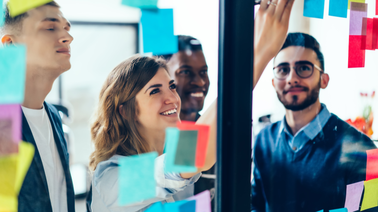 Fyra personer ser upp mot en glasvägg. En av dem sätter upp en lapp på väggen. Flera lappar i olika färger är redan uppsatta där.