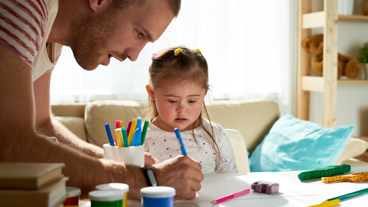 En liten tjej i prickig tröja ser förvånat på när hennes förskolelärare skriver på hennes ritpapper.
