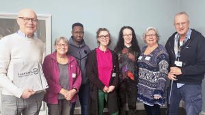 Sju personer står på rad. De ler mot kameran. Alla bär glasögon.