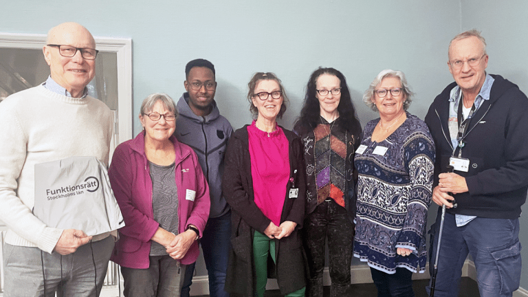 Sju personer står på rad. De ler mot kameran. Alla bär glasögon.