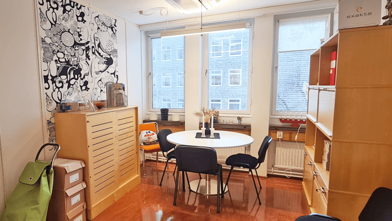 Ett rymligt kontorsrum med tre fönster. Ett förvaringsskåp står mot den vänstra väggen. I mitten står ett runt bord omgivet av tre stolar. Vid den högra väggen står en stor bokhylla.