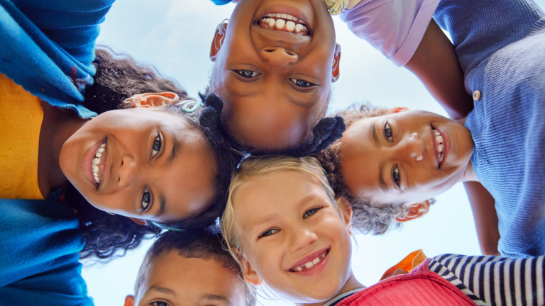 Fem barn står i en ring och håller om varandras axlar. De ler och ser ner i en kameran, som en av dem håller.
