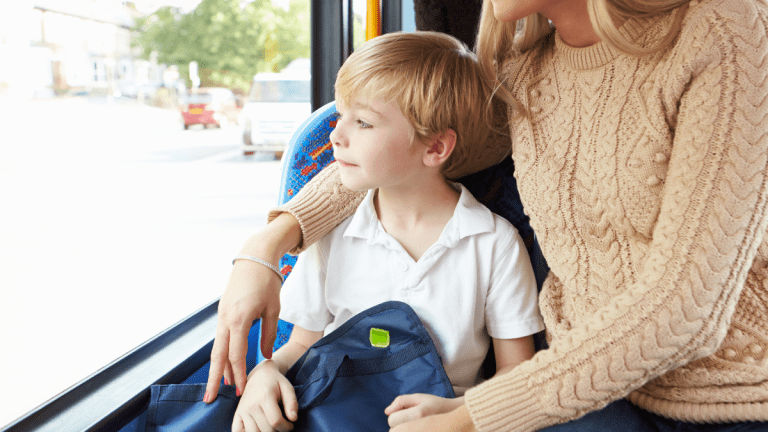En pojke med en ryggsäck i knät sitter på en buss och tittar ut genom fönstret. Hans mamma sitter på platsen bredvid honom och håller ena armen om hans axlar.