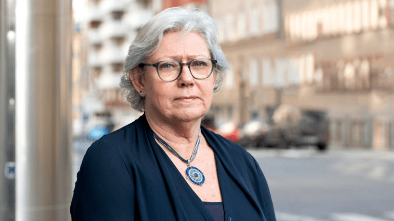 Åse ser allvarlig ut. Hon har kortklippt vitt hår och glasögon. Hon har en mörkblå tröja och ett halsband i klarblå och turkosa färger.