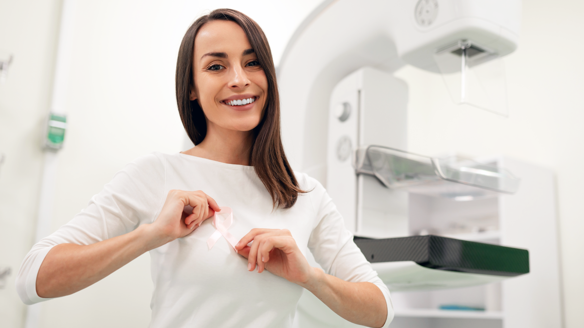 En leende kvinna håller stolt upp ett rosa band för bröstcancermedvetenhet. Hon står framför en mammografimaskin.