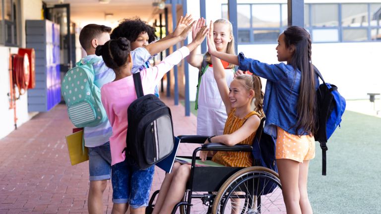 En grupp stående barn och en flicka som sitter i en rullstol har format en ring. Alla sträcker upp och ut händerna i en stor high five.