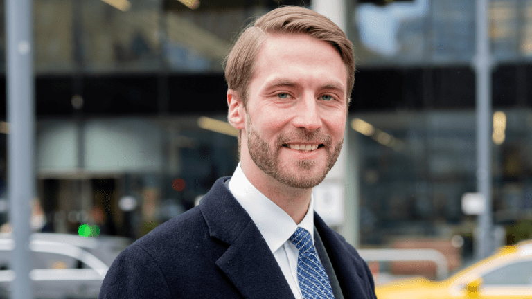 Pressbild på Moderaternas regionråd Axel Conradi. Han står utomhus klädd i kostym och slips, och ler mot kameran. I bakgrunden en byggnad med många fönster.