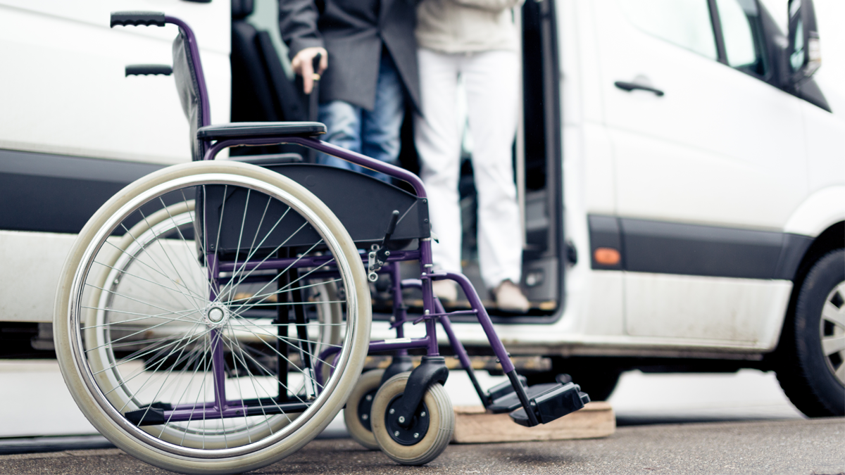 I förgrunden en rullstol. I bakgrunden får en person med käpp hjälp att komma ut ur en skåpbil.