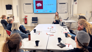 En långhårig kvinna med beige- och svartrandig blus sitter i en rullstol längst fram i en kanferenssal. Hon pratar. Bakom henne syns en presentation på en skärm. Framför henne har fyra bord ställts ihop till ett stort. Flera personer sitter runt borden och lyssnar.