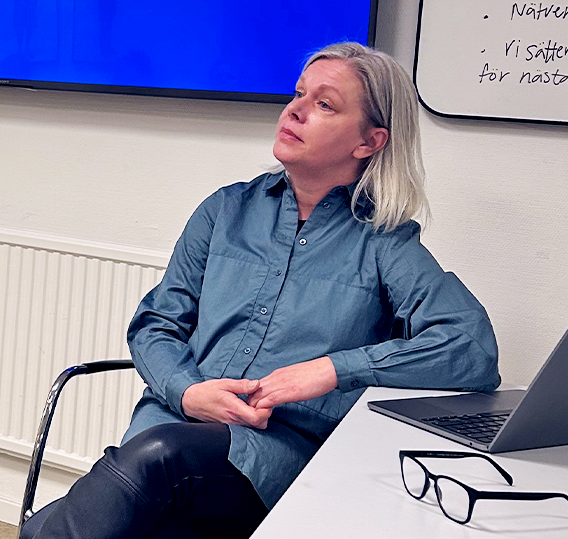 Susanna Karlsson har ljust, axellångt hår och bär en grönblå skjorta. Hon sitter längst fram i konferensrummet och i bakgrunden har hon en presentation på en skärm. Hon verkar lyssna på en deltagare i srhr-nätverket som ställer en fråga.