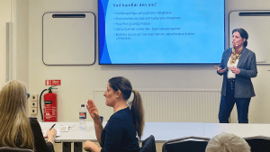 Charlotte Palmstierna från på Institutet för Mänskliga Rättigheter. Charlotte står längst fram i konferenslokalen och talar och gestikulerar med händerna. Bakom henne syns en presentation.