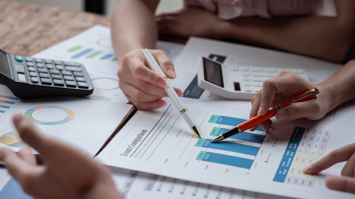 På ett bord ligger flera papper med olika diagram och tabeller. Ett par miniräknare ligger också på bordet. Två personer, troligen revisorer, pekar med sina pennor på varsin punkt i ett diagram. En annan person pekar på en tabell. En suddig hand i förgrunden gestikulerar, som att personen signalerar okunskap.
