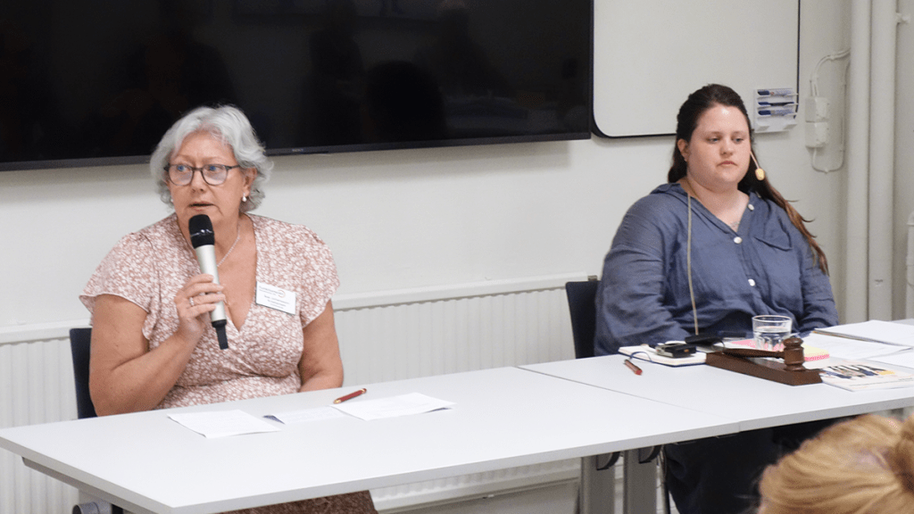 Åse har rosa- och vitmönstrad klänning, mörka glasögon och kort, vitt hår. Hon talar i en handmikrofon. Intill henne sitter Mikaela, som har långt, brunt hår och en blå skjorta.