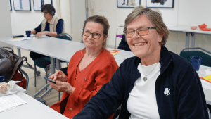 Två kvinnor, båda i glasögon. Den ena är klädd i orange och har uppsatt, brunt hår. Den andra har en blå jacka över en vit tröja. Båda ler stort mot kameran.