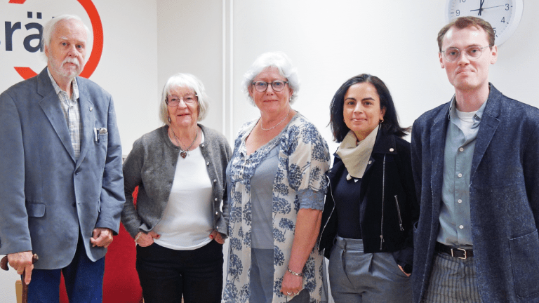 Fem personer står på rad framför vår logga på väggen.