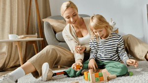 En blond kvinna sitter på en fluffig matta på golvet. Framför henne sitter hennes barn. Barnet har en benprotes. De leker med klossar och båda ser glada ut.