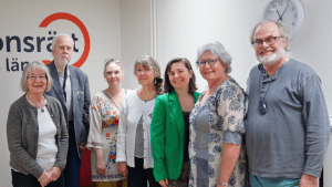 Sju personer står på rad framför vår logga på väggen. Alla ler mot kameran.