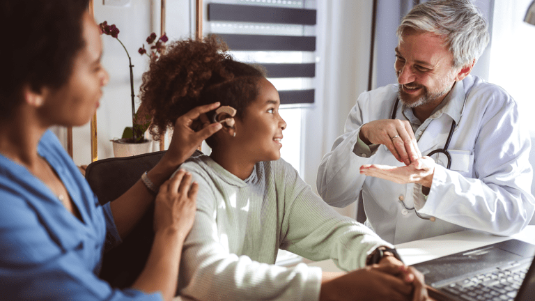 En läkare i vit rock tecknar något till en flicka. Båda ler. Bakom flickan skymtar en blåklädd sjuksköterska, som justerar en hörapparat på flickans ena öra. Många barn med funktionsnedsättning har erfarenhet av både hjälpmedel och övergångsvård!