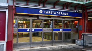 En entré, med en skylt ovanför som upplyser om att man nått Farsta strands tunnelbanestation. Genom de stora glasrutorna i två par metalldörrar skymtar spärrarna och en butik.