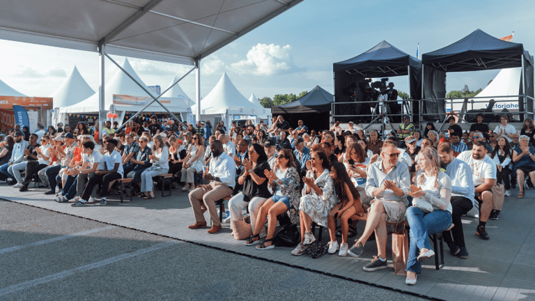 Flera rader av fullsatta träbänkar, där en publik sitter och applåderar. Solen ligger på, trots att det finns ett tak uppsatt ovanför. I bakgrunden tält med kamera- och ljudutrustning.