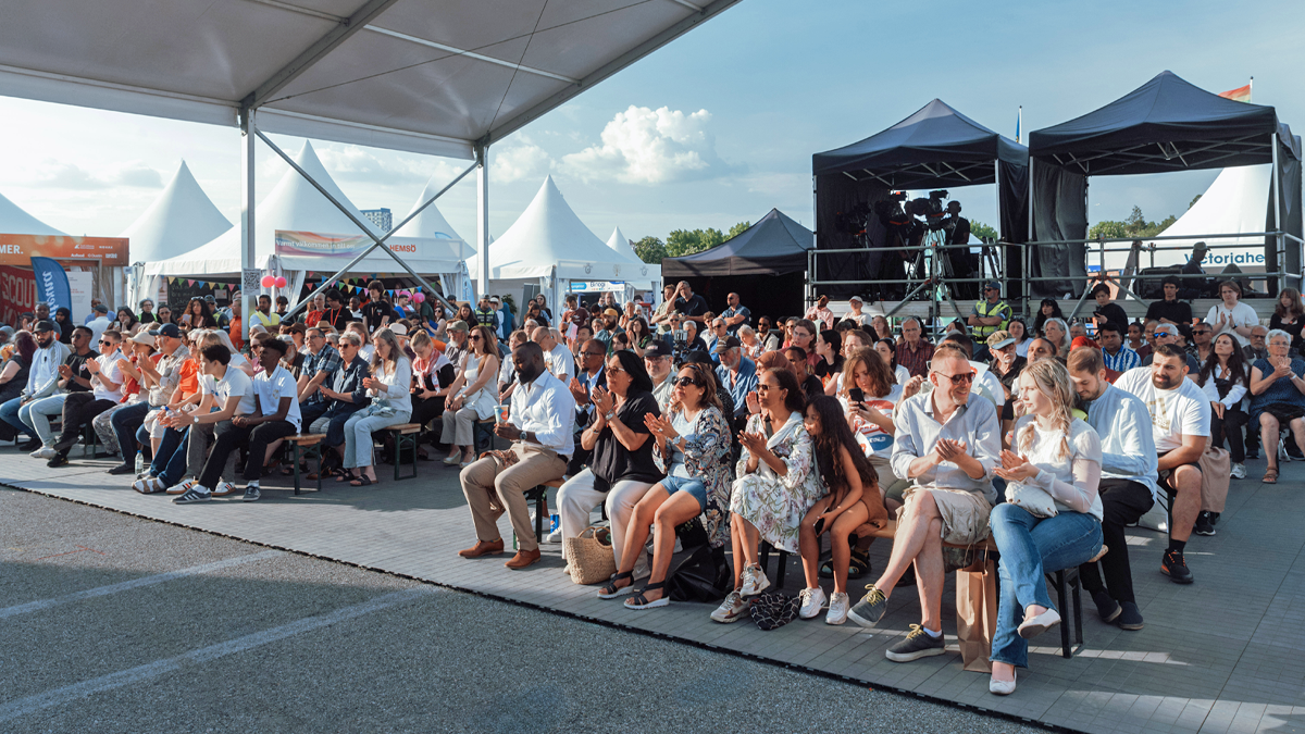 Flera rader av fullsatta träbänkar, där en publik sitter och applåderar. Solen ligger på, trots att det finns ett tak uppsatt ovanför. I bakgrunden tält med kamera- och ljudutrustning.