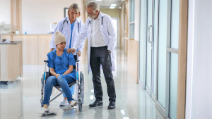 En kvinna i rullstol i en sjukhuskorridor. En sjuksköterska bakom henne håller i rullstolens handtag. En läkare står intill och håller en hand på kvinnans axel.