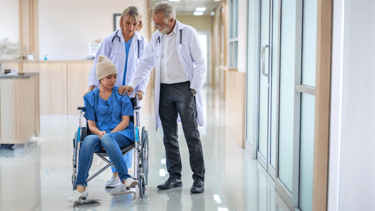 En kvinna i rullstol i en sjukhuskorridor. En sjuksköterska bakom henne håller i rullstolens handtag. En läkare står intill och håller en hand på kvinnans axel.