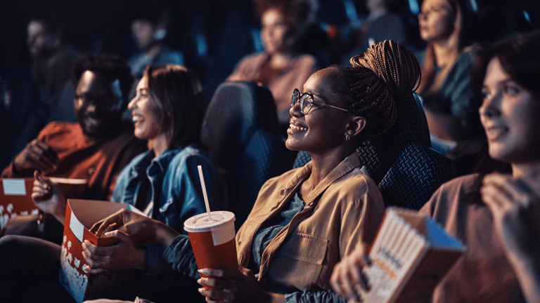 Flera personer i en biopublik. Många håller en popcorn-kartong. Alla tittar mot skärmen och ler.
