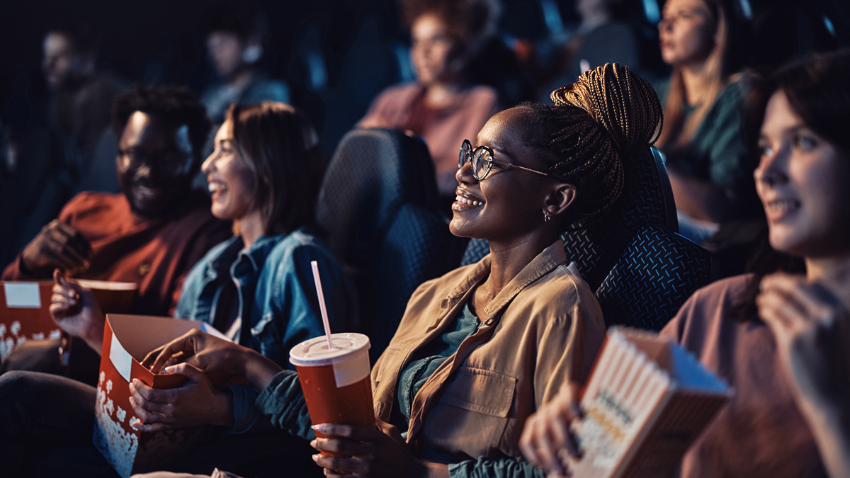 Flera personer i en biopublik. Många håller en popcorn-kartong. Alla tittar mot skärmen och ler.