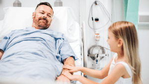 En man ligger i en sjukhussäng. Har bär en ljusblå sjukhussärk och han har plastslangar i näsan. Han ler mot en liten flicka som står intill sjukhussängen. Flickan, som har långt, blont hår och bär ett vitt linne, ler tillbaka. Hon håller händerna på mannens arm. Barn som anhöriga.