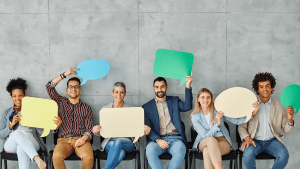 Flera personer sitter på rad. Alla ler och håller upp varsin utklippt pratbubbla med olika färger och former. Bubblorna representerar deras olika åsikter. En skicklig opinionsbildare kan påverka samhällsdebatten.