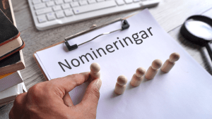 Ett vitt papper med texten "nomineringar". En rad små träfigurer står på pappret. En hand håller i ännu en träfigur och ställer den på pappret med de andra figurerna.