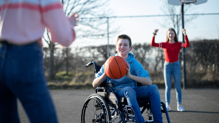 En ung kille är fokus. Han sitter leende i en rullstol och är just på väg att kasta en basketboll. Resten av bilden är suddig och ur fokus. Bakom killen står en tjej framför en basketkorg och håller upp armarna. I förgrunden står en annan tjej vänd från kameran mot killen. Hon håller upp armarna för att fånga bollen.