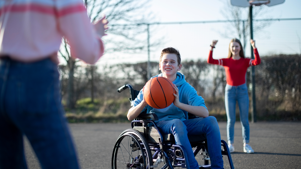 En ung kille är fokus. Han sitter leende i en rullstol och är just på väg att kasta en basketboll. Resten av bilden är suddig och ur fokus. Bakom killen står en tjej framför en basketkorg och håller upp armarna. I förgrunden står en annan tjej vänd från kameran mot killen. Hon håller upp armarna för att fånga bollen.