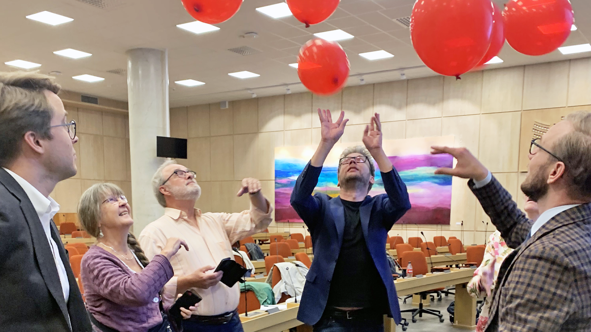 Fler personer står i en halvcirkel i landstingssalen. Flera håller i sina telefoner och sträcker samtidigt upp sina händer för att hålla ett flertal ballonger i luften.