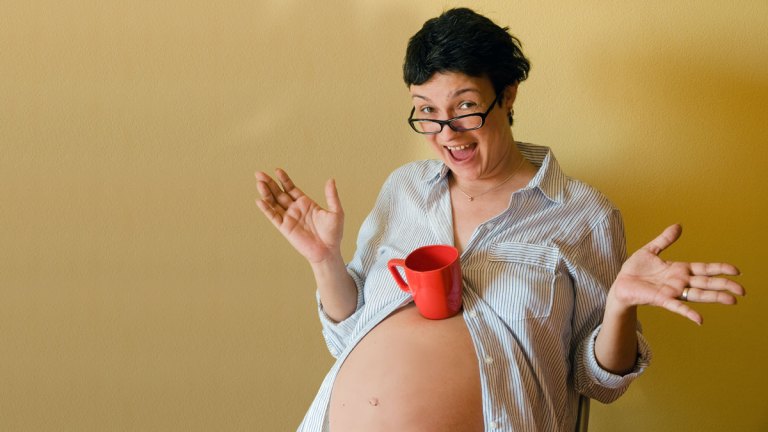 En leende kvinna balanserar en kaffemugg på sin stora gravidmage.