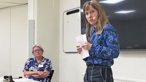 Marian har en blåmönstrad skjorta och utsläppt, brunt hår. På huvudet bär hon en mikrofon, som hon talar i. I bakgrunden sitter ordföranden Åse. Åse bär en blå klänning med vitt mönster, och hon tittar fokuserat på Marian.