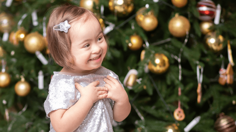 En liten flicka i silverglittrig klänning står framför en julgran. Hon ler stort.