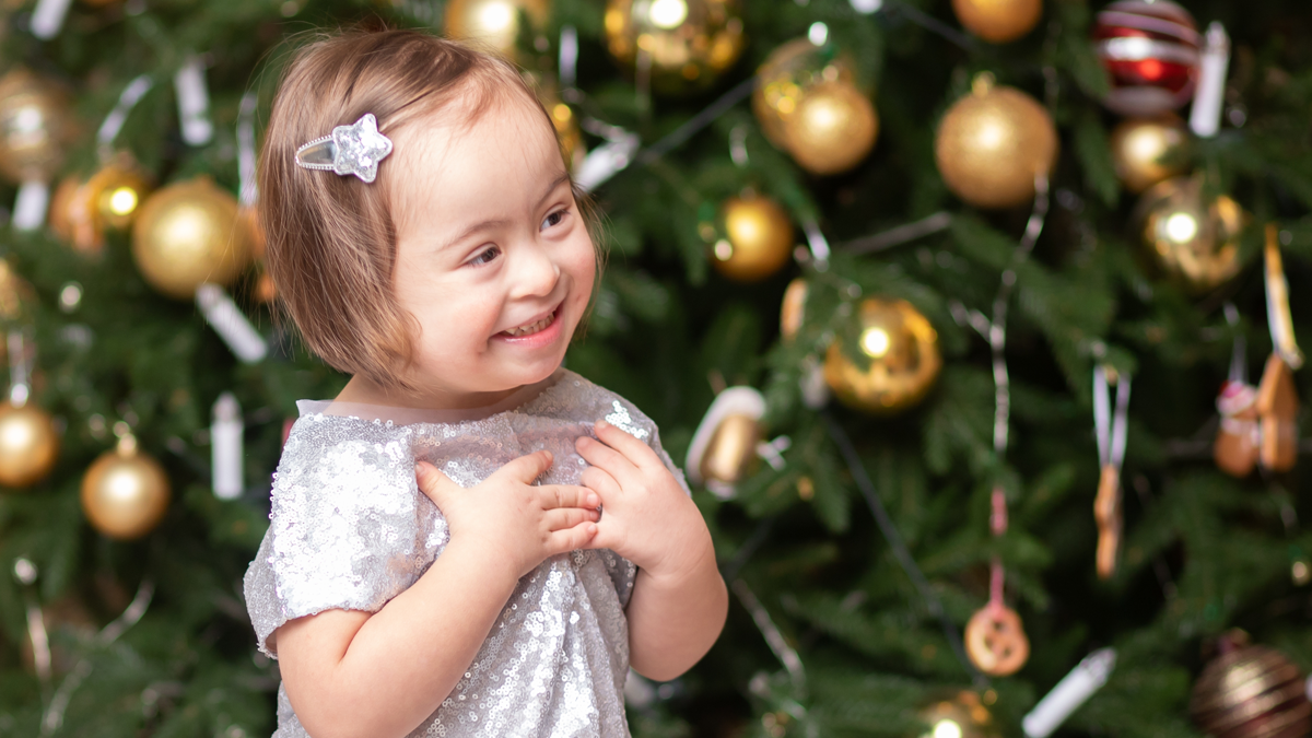 En liten flicka i silverglittrig klänning står framför en julgran. Hon ler stort.