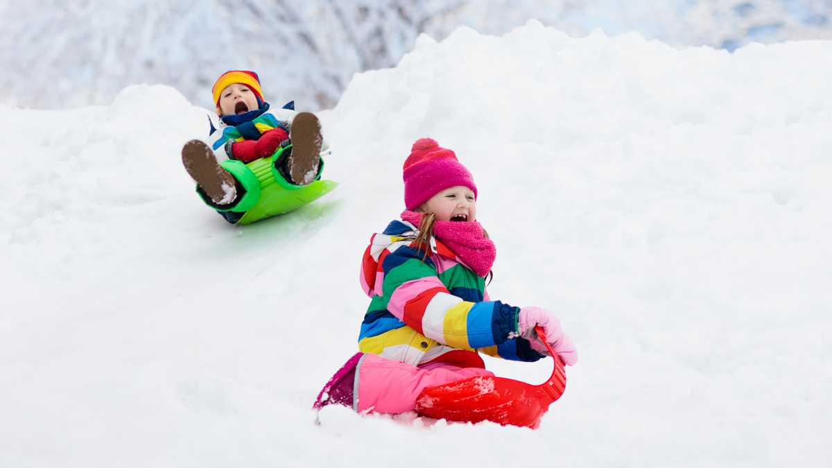 Två glada barn i färgglada kläder åker pulka nerför en backe.