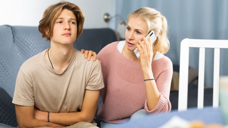 En tonårskille håller sig om magen. Hans mamma sitter bredvid och talar i sin telefon. Båda ser oroliga ut.