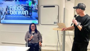 En asiatisk kvinna i rullstol håller i en handmikrofon och ler. Intill står hennes kollega, en vit man, vänd mot projektorskärmen i bakgrunden, och talar i en annan handmikrofon.