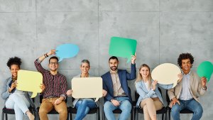 Flera personer sitter på rad. Alla ler och håller upp varsin utklippt pratbubbla med olika färger och former.
