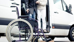 En sjuksköterska hjälper en man med käpp att ta sig ut ur en färdtjänstbil. I förgrunden står en rullstol.