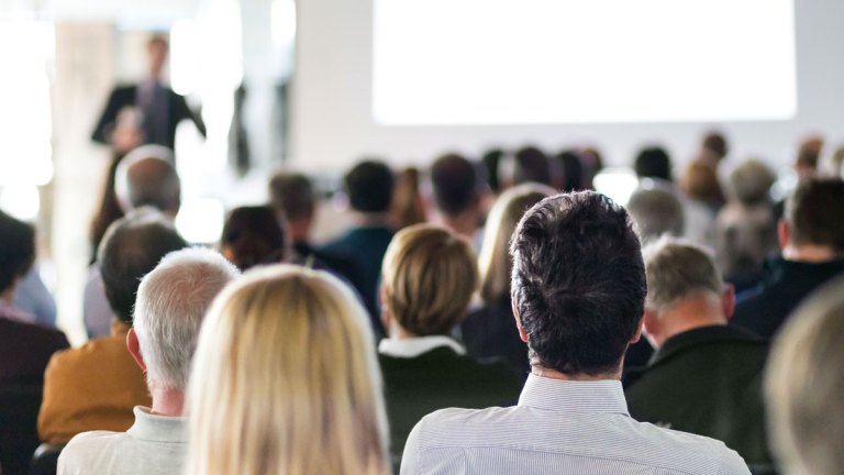 Flera personer sitter i en konferenssal och lyssnar på personen längst fram, som pratar i en mikrofon.